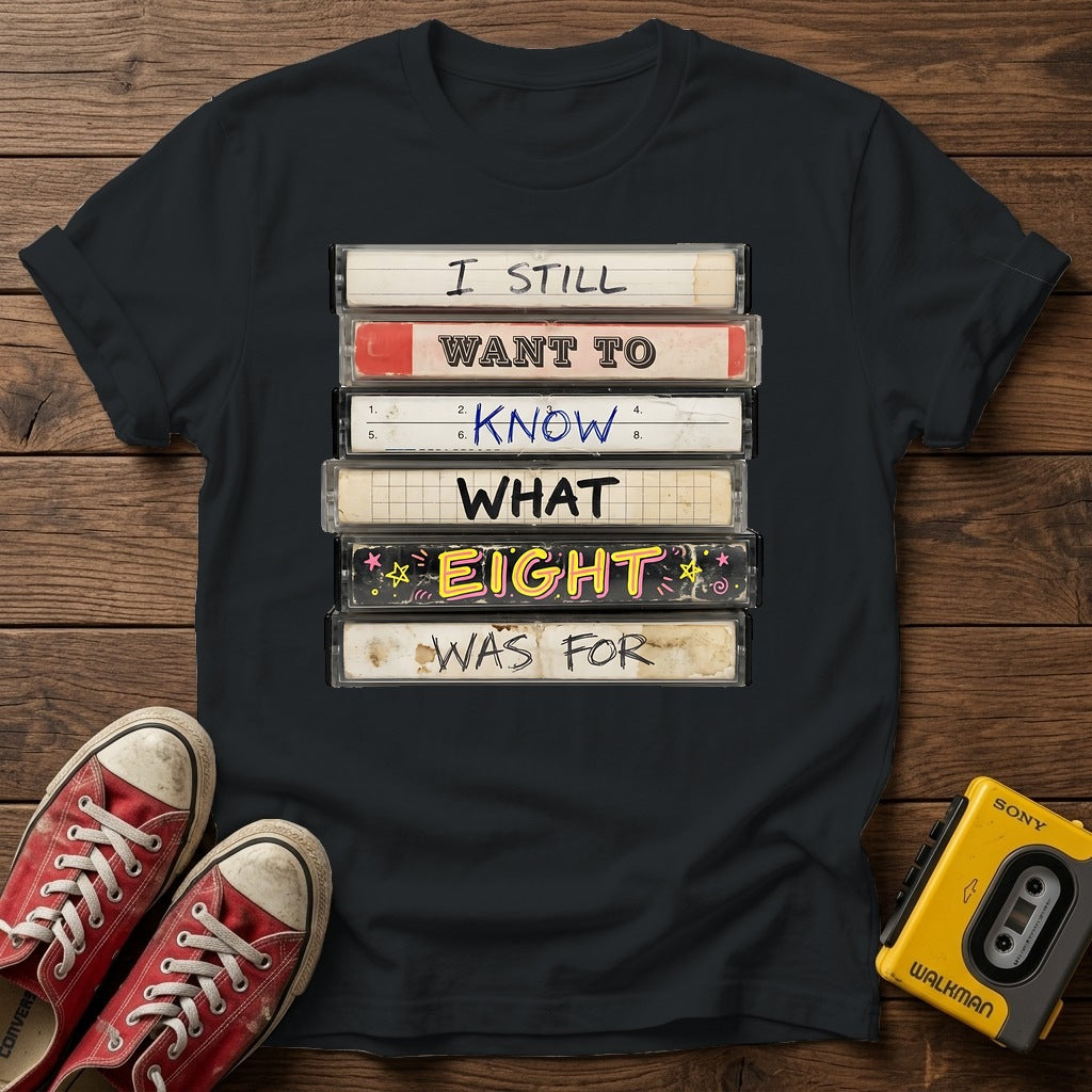“I Still Want To Know What Eight Was For” Retro VHS Stack T Shirt — Black wood flat-lay