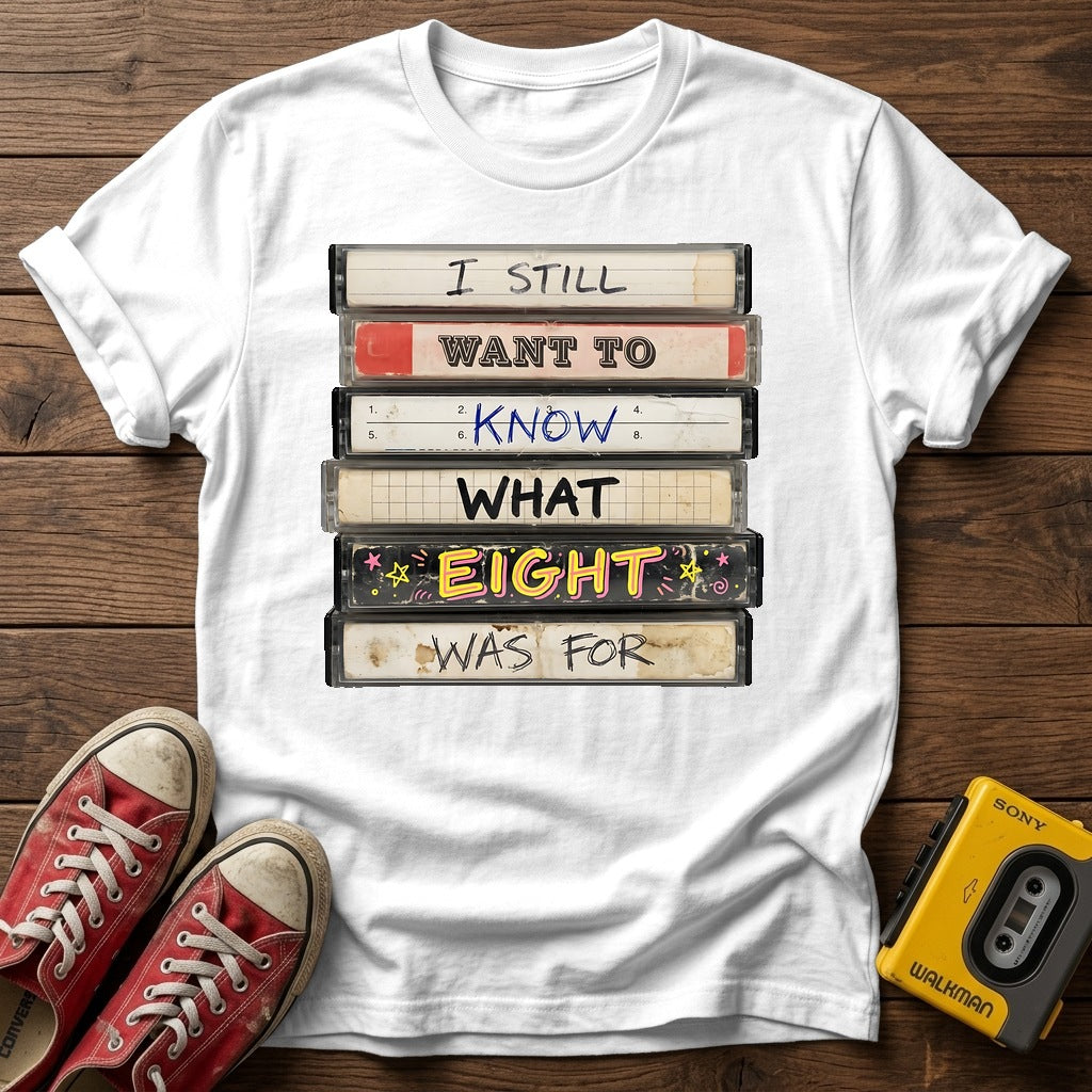 “I Still Want To Know What Eight Was For” Retro VHS Stack T Shirt — White wood flat-lay